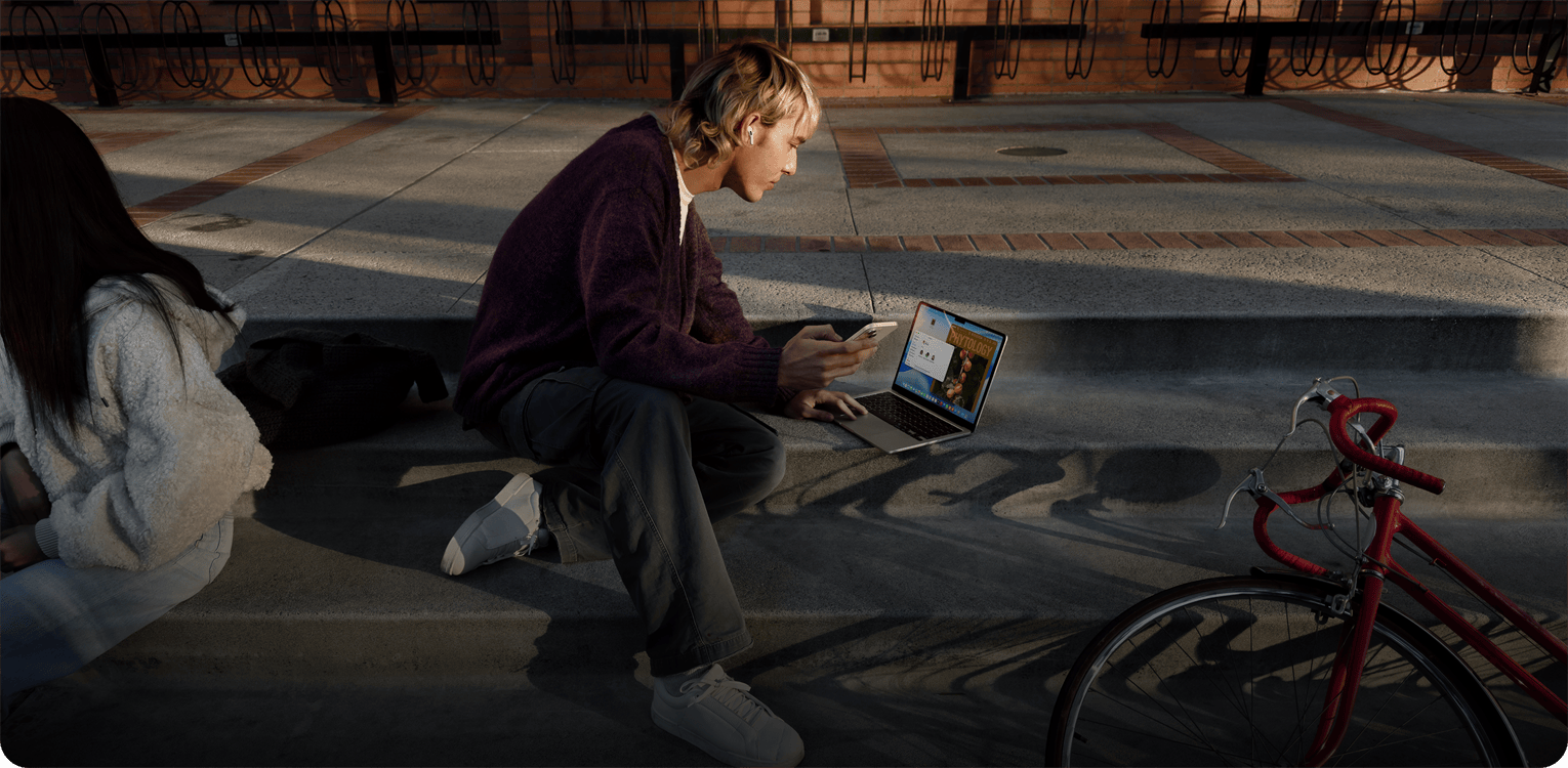 Osoba, která sedí venku na betonových schodech, má nasazené jedno sluchátko AirPodů, drží iPhone a pomocí trackpadu ovládá MacBook položený na zemi před sebou, poblíž leží červené jízdní kolo a sedí druhá osoba