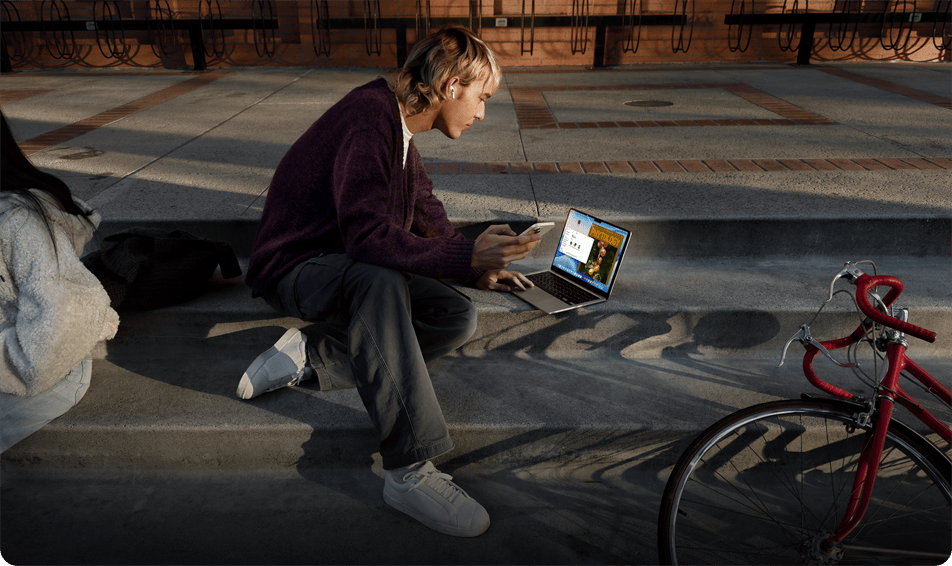Osoba, která sedí venku na betonových schodech, má nasazené jedno sluchátko AirPodů, drží iPhone a pomocí trackpadu ovládá MacBook položený na zemi před sebou, poblíž leží červené jízdní kolo a sedí druhá osoba